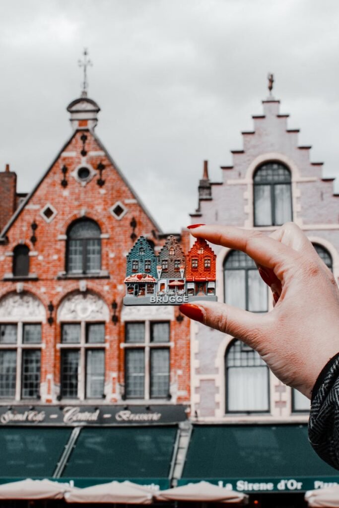 A hand holding a miniature Bruges building with traditional Belgian architecture in the background.